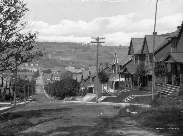 A view of south Sixth Avenue, looking down hill. Houses line both sides of the street, with one tree visible in the foreground and multiple trees lining both sides of the streets in the middle ground. Farmland or unoccupied, non-residential land is in the background.