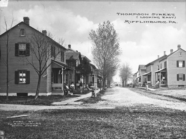 A view of Thompson Street, with people sitting on their porches and standing in the street. Trees line the street. Caption reads: "Thompson Street, (looking east), Mifflinburg, PA."