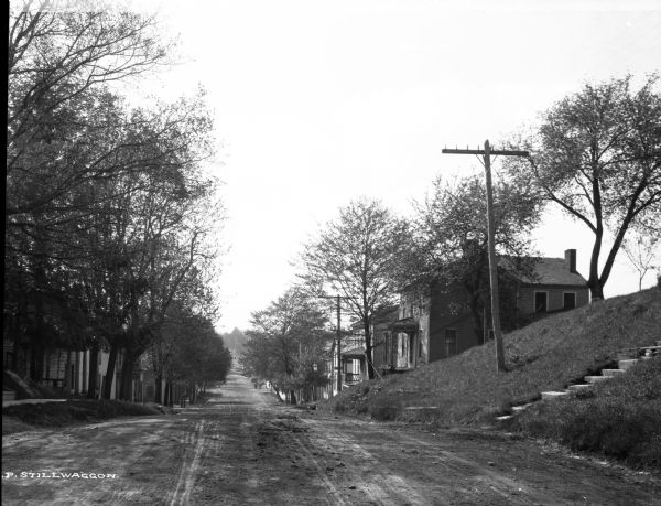A view of Main Street. Houses line both sides of the street, as do trees, which partially obscure the houses. Telephone and/or electric lines run parallel to the street on the right side.