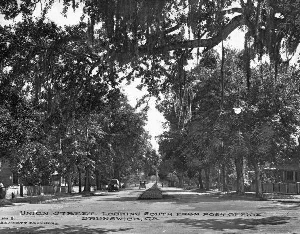 A view of Union Street, which is a wide thoroughfare with a grass median. On the left side of the median is both a car and truck. On the right side a figure walks or rides a bicycle. Trees line the lane on both sides, partially obscuring the houses. Caption reads: "Union Street, looking south from Post Office, Brunswick, GA."