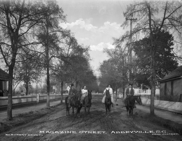 A view of Magazine Street, with four people, two men and two women, posing on their horses. Trees line the street, and houses and fences are on both sides. Caption reads: "Magazine Street, Abbeyville, S.C."