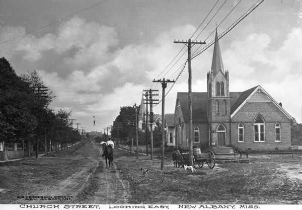 A view of Church Street. Trees line both sides of the street, with telephone and/or electricity poles. The trees obscure the buildings to the left, and two churches are on the right. In the foreground, a dog stands next to a wagon, and a man on horseback rides down the street. Caption reads: "Church Street, Looking East, New Albany, Miss."