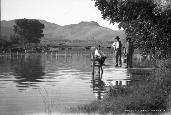 A view of three men, one sitting and two standing, on a lake dock. In the background on the far shoreline are cattle grazing. Foothills are in the background.  