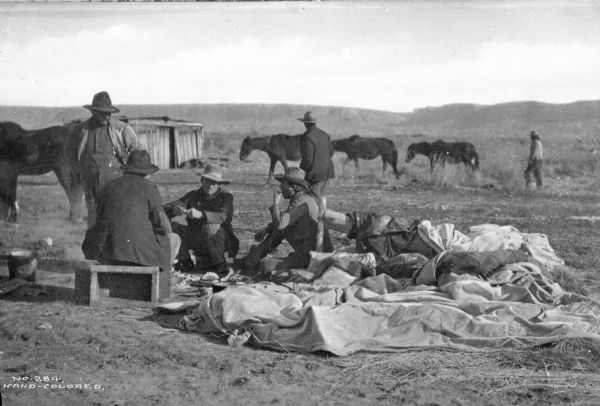 Four ranch hands sitting and standing on the left. In the background two men walk near horses.