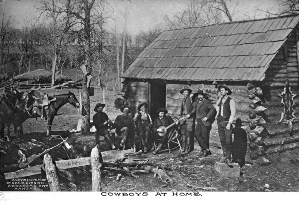 A view of cowboys posing outside of their log cabin dwelling. Three stand against the wall, while four sit. The sitter on the right has a banjo. In the left foreground is a wood pile, and a horse lingers behind it. Caption reads: "Cowboys at Home."