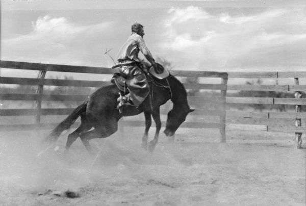 A cowboy holding his hat in his hand is riding on a bucking horse in a fenced enclosure. The horse is kicking up dust.