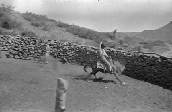 Slightly elevated view of a ranch hand riding a bucking horse. To the left is a man or boy standing near the stone wall enclosure. In the background are hills and vegetation.