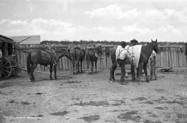 Three ranch hands saddling and packing a horse in an enclosed fence area. One man has his foot pushing against the horse while tightening the rope. Three other horses, already saddled, are standing on the left near a wagon and outbuilding. In the background are trees and shrubs.