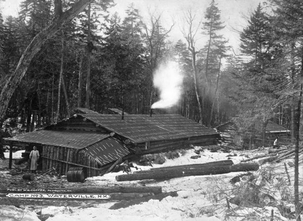 Elevated view from hill toward two buildings at the lumber camp. A man stands outside of the structure nearest the camera. In the foreground are logs, with trees in the background surrounding the camp. Caption reads: "Camp No. 3, Waterville, N.H."
