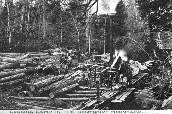 Slightly elevated view of a logging camp, with a dozen or so men posing by the wagons, piles of logs and machinery. There are both logs and milled lumber. Surrounding the scene and in the background is the forest. Caption reads: "Logging Camp in the Kentucky Mounains."