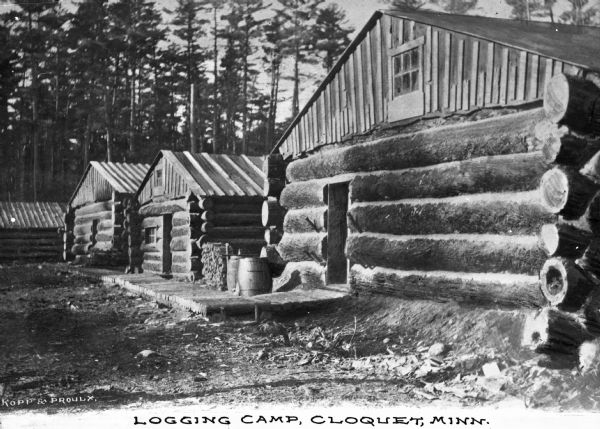 A view of the log cabin housing at a logging camp. Caption reads: "Logging Camp, Cloquet, Minn."