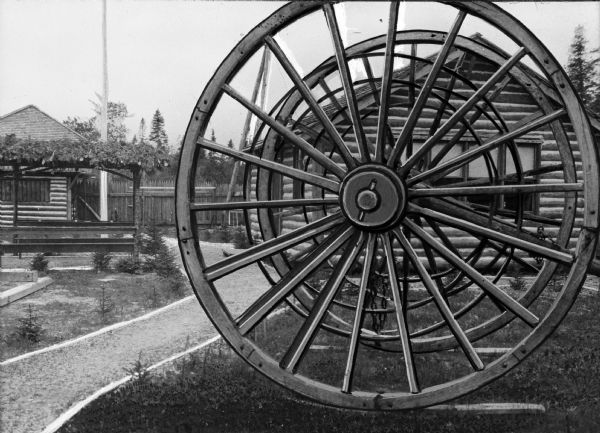Two large logging wheels outside of Fort Algonquin, located off of US 21. The wheels sit to the right of a path, and in front of a small log structure. In the background, to the left of the path, is a pergola and similar log structure.
