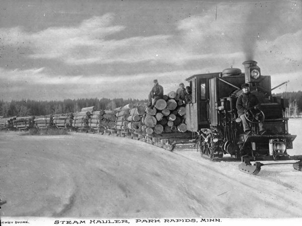 A view of a steam hauler pulling logs across ice on sleds. Four men are posing, with one man sticking his head out of  a window of the hauler; one man is sitting in front behind the driver's wheel; and two men are sitting behind the hauler on the logs. A forest isn in the distant background. Caption reads: "Steam Hauler, Park Rapids, Minn."