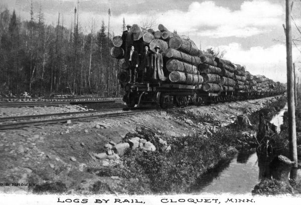 Three men are standing on the last railroad car which is carrying logs down a railroad track. Another set of tracks are running parallel on the left, and behind that is a forest. In the foreground on the right is a ditch and stream, also running parallel with the tracks. Caption reads: "Logs by Rail.  Cloquet, Minn."