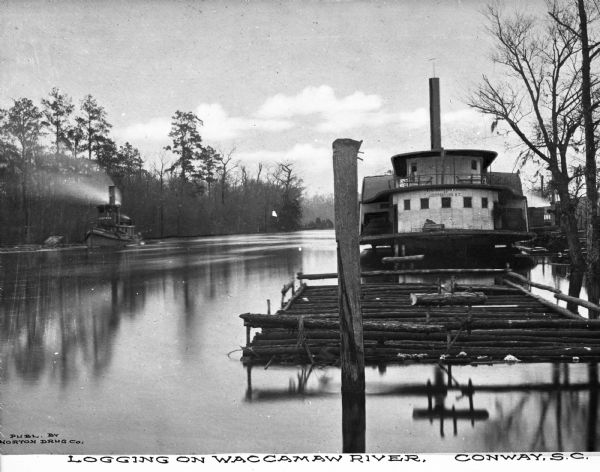 Logging scene on Waccamaw River. On the right is a group of logs floating  in a contained area in front of a boat. In the background on the left is another boat moving along the shoreline. Caption reads: "Logging on Waccamaw River, Conway, S.C."