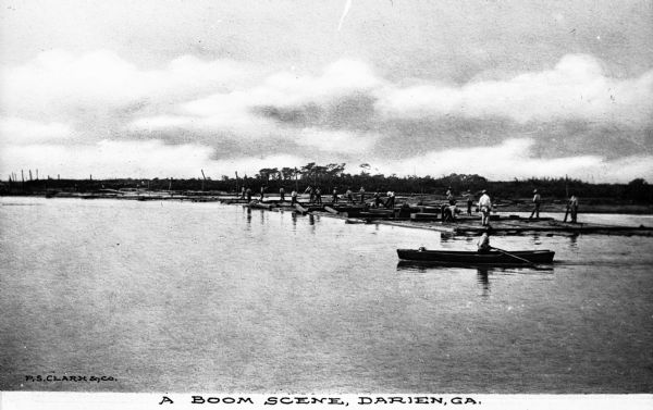 View across water toward men standing and working on logs in the middle of the river. Booms help to collect the logs that are shipped down river from the forest. In the foreground is a man a rowboat. Caption reads: "A Boom Scene, Darien, GA."