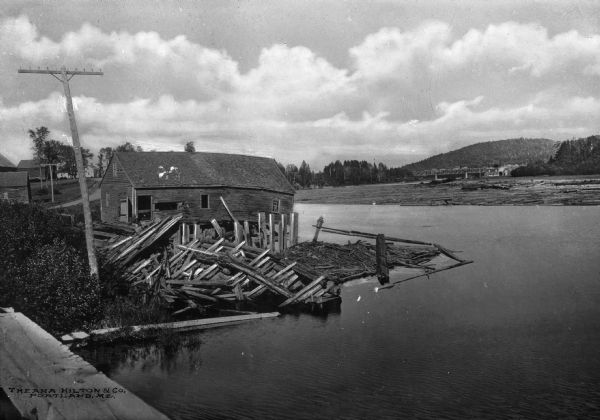 Elevated view across water toward mill ruins on the shoreline. A pile of logs are next to the dilapidated structure. In the far background is a bridge and other structures, wit hills looming behind.
