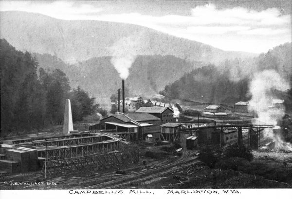 Elevated view of Campbell's Mill, which specialized in lumber. Situated in the midst of the forest are smokestacks, railroad tracks, and several buildings. In the left foreground are above-ground tracks on which to move the finished lumber. Caption reads: "Campbell's Mill, Marlinton, W.Va."