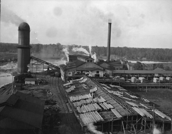 Elevated view of the Brooks Scanlon Company, which specialized in lumber. The complex sits on a river. The lumber is lying out on the tops of roofs and in piles in the foreground. A tower is next to the river on the left, and there are various outbuildings that extend from the foreground to the middle ground. Smoke rises from a number of chimneys as well as a tall smokestack. Forests are in the background on the other side of the river.