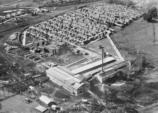 Aerial view of the Baldwin Lumber Company, which sits next to a river. The main building consists of two long, rectangular structures that sit perpendicular to each other, and there are railroad tracks that run up to the intersection of the structures. Beyond the buildings is an area filled with large stacks of finished lumber. Small buildings are in the distance.