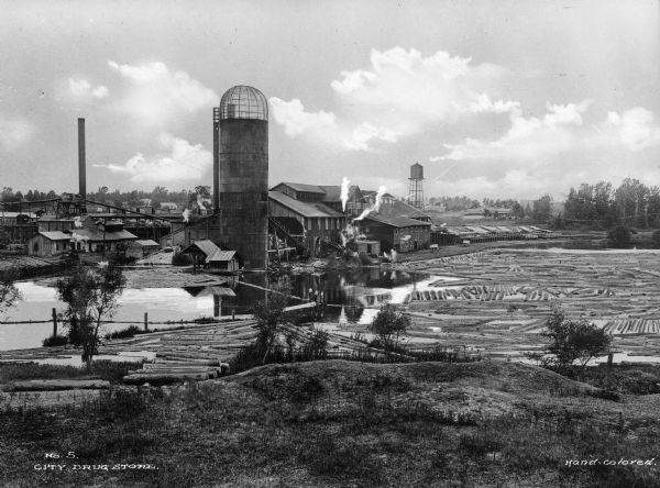 View looking down hill toward the sawmill and logs floating in the pond. Company outbuildings are on the left, with a smokestack and silo. A water tower is in the background.