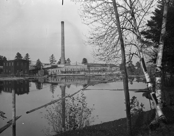 A view of a Grand Rapids lumber mill.  In the foreground is a body of water, with logs floating in it. In the center of the image is a smokestack, which reflects in the water.  A main, light-colored stone  building stands below the stack.