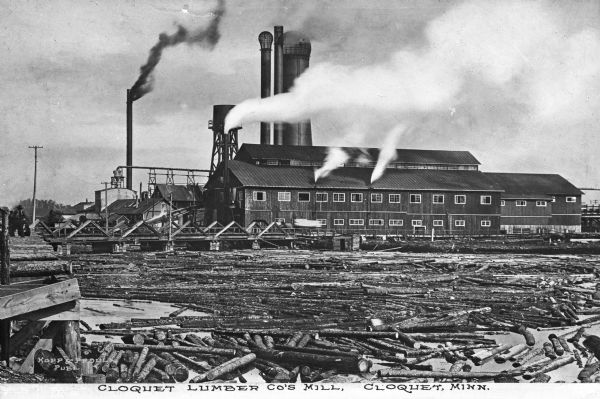 View across water covered with logs toward the mill. Smoke and steam is coming from a smokestack and chimneys. The mill building has smaller outbuildings connected to it. Caption reads: "Cloquet Lumber Co's Mill, Cloquet, Minn."