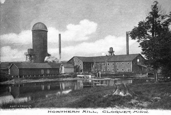 View from shoreline across water toward the mill. To the right of the main building are two smaller structures, with a watertower and silo behind. Two smokestacks are in the background. The main structure has diamond-shaped windows and open skylights on the roof. Caption reads: "Northern Mill, Cloquet, Minn."