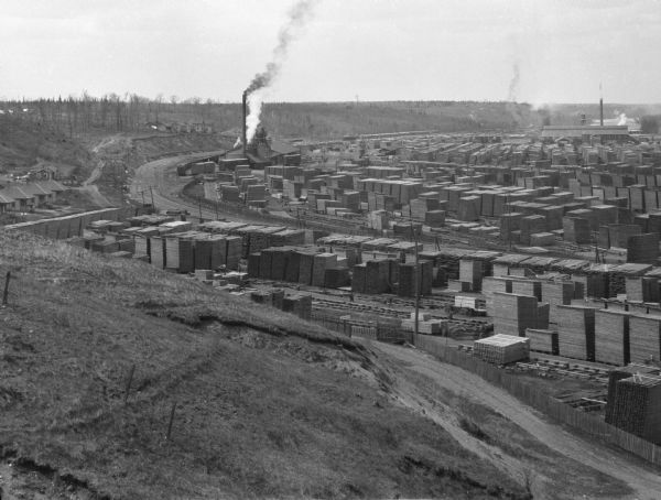 Elevated view from hill toward a lumberyard, with small workers' homes on the left. A dirt path and wooden fence surround the lumberyard, which is filled with stacks of lumber. Railroad tracks are around the yard. A small grouping of outbuildings and the main complex, with silo, smokestack, and chimneys are in the background.