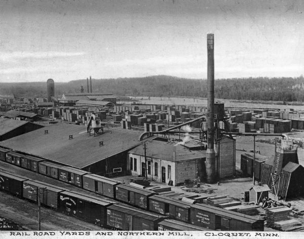 Elevated view of the railroad tracks, lumberyard, and mill of Northern Lumber Mill. In the foreground is a long line of railroad cars sitting on the tracks in front of buildings. Stacks of lumber and the mill are in the background. Caption reads: "Rail Road Yards and Nothern Lumber Mill, Cloquet, Minn."