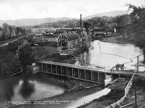 Elevated view of the mill, dam, and railroad tracks, and forests surrounding the area. The lumber mill itself, with main building complex and smokestack is in the background. In front of this is a barn-like structure with a pointed arch gable. In the foreground is a river, and spanning it is a combination bridge and dam. Caption reads: "Lumber Mill and Water Power at Bonner's Ferry, Idaho."