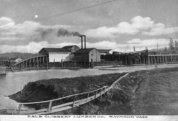 View toward the lumber mill. The mill complex is on the far shoreline, with two smokestacks. The building on the left side  has a track the extends from the structure to the river, which is in front of the mill.  Behind the main complex are other buildings, with a lumberyard on stilts, with neatly-arranged lumber piles. Caption reads: "Kalb Glibbert Lumber Co., Raymond, Wash."