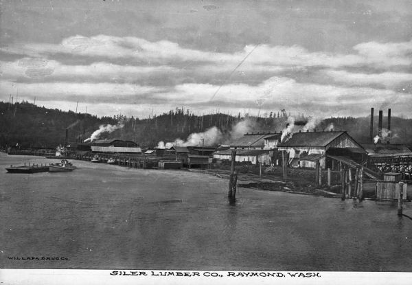 Elevated view across water toward the lumber mill. On the left is a boat, moving away from the loading dock. Smoke and steam emanate from various building complexes. Behind the structures, on the right, are three smokestacks. Caption reads: "Siler Lumber Co., Raymond, Wash."

