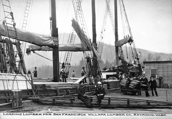 View of lumber being loaded onto a boat with men on the boat and on the dock. On the dock is a small building, with a group of men, two in suits, leaning against a pile of lumber waiting to be loaded. In front of them are two men sitting on a wooden structure, and above them are sails and rope ladders on the ship. Caption reads: "Loading Lumber for San Francisco, Willapa Lumber Co., Raymond, Wash."