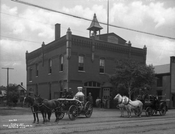 A view of two horse-drawn engines sitting in front of the fire station.  The engine on the left holds the water tank and is pulled by two horses.  Two men ride up front while a third stands on the back of the wagon.  The second engine, to the right, has men posed in the front and one in back, standing. Four men stand by the fire station doors. Electric lines run across the image, and tracks line the brick street.  Printed in the bottom left corner: "No. 45 Publ. by the Long & Taylor Co Akron, Ohio."