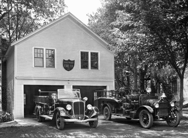 A view of the fire station and two fire trucks, parked in front of garage doors.  Centered between them is a shield-shaped sign the says "Hayward Fire Co."A vine-covered building sits to the right of the station.  Trees grow on both sides of the structure.