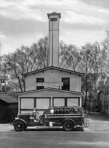 A view of the fire department and engine.  The station is wooden, with a look-out tower.  It is small, and surrounded by other wooden structures, mostly homes.  A sign says "Bethel Fire Dept. No 1."  The engine is parked in front of the station.