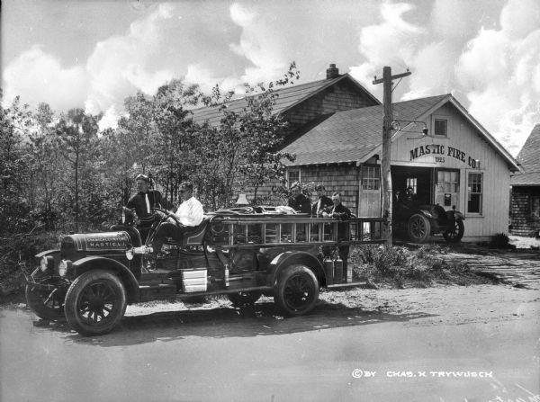A view of the Mastic, Long Island firemen. A fire engine is parked on the street, to the left of the station. Five firemen are posing in it, two in the front and three in the back. Another engine is sitting halfway outside of the building, in the driveway, with two men in it. The fire station is covered in shingles, with a large, two-story section behind a one-and-a-half story garage in front, with the facade covered in vertical wood siding. Centered above the door and window is "Mastic Fire Co.," with "1925," painted below. A street light is hanging over the driveway, and a shingle-covered building is visible to the right. Printed in the bottom right corner is a copyright symbol and the words: "By Chas. H. Trywusch."
