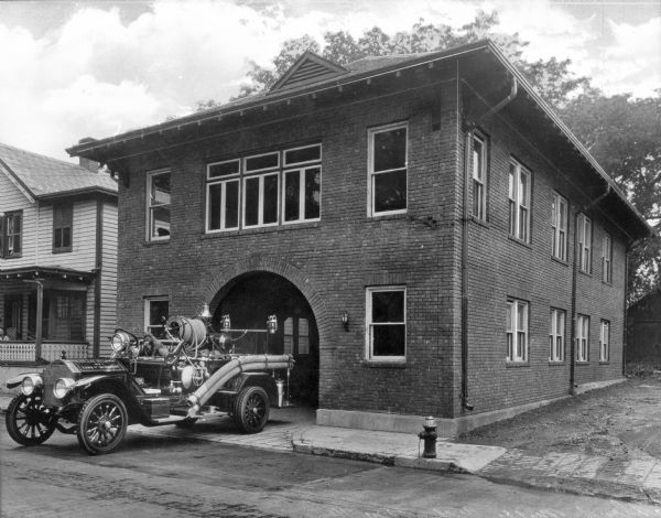 A view of the building and engine.  The building, made of brick, contains a garage door opening in an arch shape, accented by the brickwork around the arch.  The foundation is possibly made of concrete, and the roof is hipped.  Two downspouts are visible on the right side of the building. A house sits to the left of the stations, which is fronted by a paved sidewalk. The engine, with a pudgy dog sitting in the front seat, sits outside of the station, half in the driveway and half in the middle of the street.