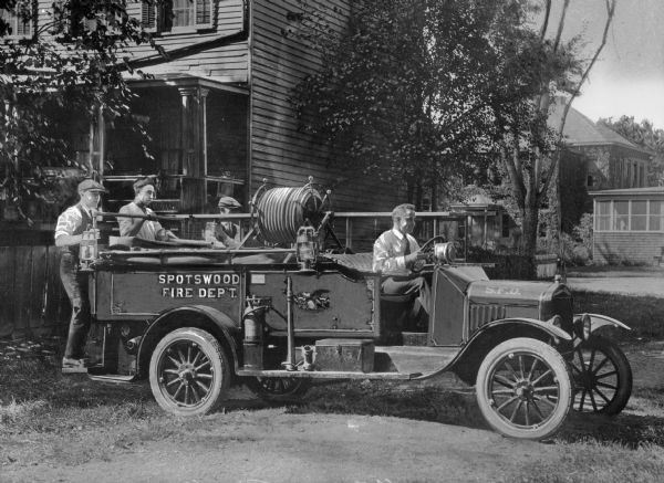 A view of the engine, with four men posing on it, one in front and three in the back.  It is parked in an empty lot, with a house to the left.  This structure has clapboard siding.