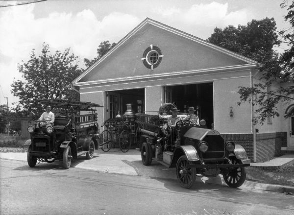 A view of the fire department and its engines.  The garage consists of two bays, with a small, circular window placed above, the gabled roof forming a triangular pediment.  The deeply-set window is multi-paned with brick outlining.  A door is placed on the right side, set back from the front.  The engines sit partially in the driveway and partially in the street, with the left engine carrying an attachment cart that appears to contain the water tank.  Only one man appears in this engine, while the right on carries two men, both in the front seat.