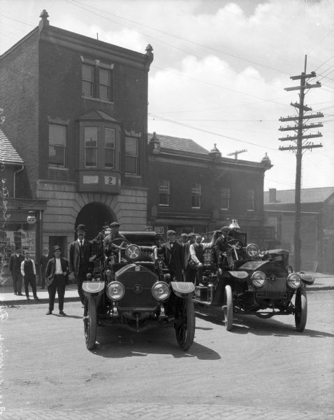 A view of the fire department and two engines in the middle of the street.  The engine on the left contains three men, one at the wheel and two standing on both sides.  The right one contains five men, one at the wheel and the others standing on the left side. The building itself is surrounded on both sides by other businesses and shop fronts, the business on the left being the tailoring business of Louis Leonardo, seen on the window and light fixture.