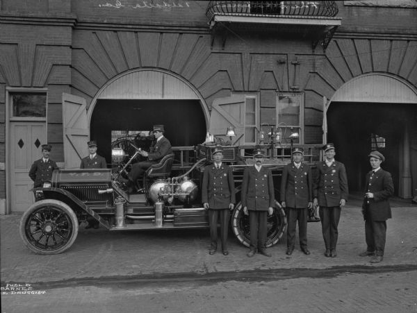 A portrait of the firemen, standing in front of the engine. The engine is seen from the side, and eight men, in uniform pose, five in front of the engine, one at the wheel, and two to the left.  Printed in the bottom left corner: "Publ. by Barnes [?]e Druggist."