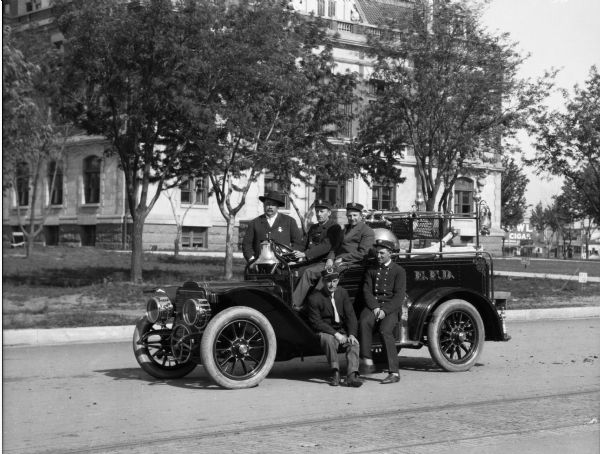 A portrait of the firemen posed with the engine, parked in the middle of the street. The five men are grouped closely together, two sitting on the side of the engine, two in the front seat, and one stands behind the engine, near the front.