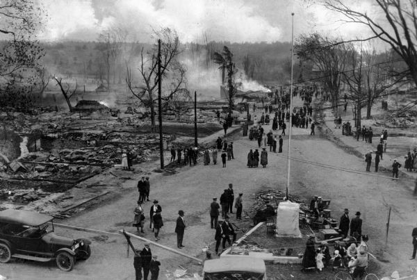 Overhead view of a crowd, consisting of men, women, and children standing among what appears to be charred ruins. Smoke is rising in the background.