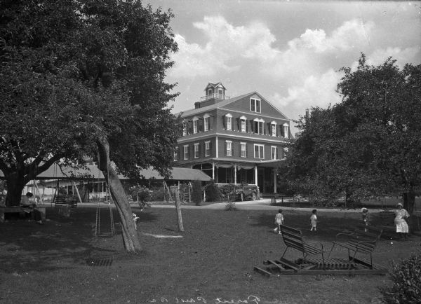 View across lawn toward the Unity House. On the left are two women reading in swings fastened to a large tree. A covered walkway behind them leads to the house, which has a wraparound covered veranda. In the right foreground, three children play on the lawn near a woman sitting in a swing. Two sliding benches are facing each other in the foreground.