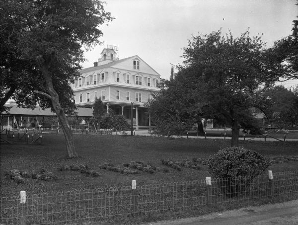 View over fence and lawn toward the Unity House. Four women are sitting in the covered walkway that leads to the covered, wraparound veranda.  There are trees in the yard and shrubs near the fences, and small plants have been grouped together to form the word "Unity" in the foreground near the fence. There is a driveway on the right.