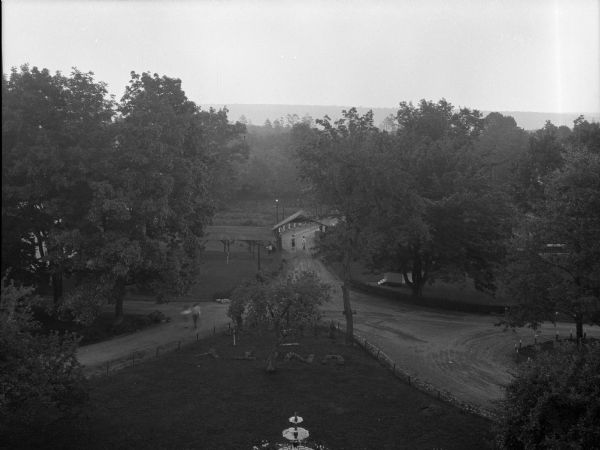 Elevated view of the lawn and driveway, with a fountain in the foreground, and plantings that spell "Unity." Two women are walking on the left path, and in the center are two women who are passing beneath the wooden covered entrance in the background. A woman is holding a baby in the covered pathway.
