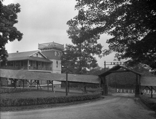 View toward the covered entrance and covered pathway. Above the roof is sign that reads, in reverse: "Welcome." To the right is a large structure, probably rooms or apartments, which has a square tower-like structure on the corner.