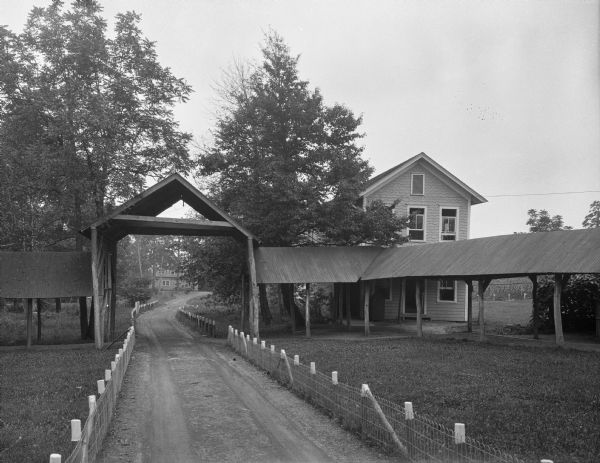 View down fence-lined road towards the covered entrance and walkway. A two-story house is in the background on the right. A fence is along the dirt path, with a small building in the far background.